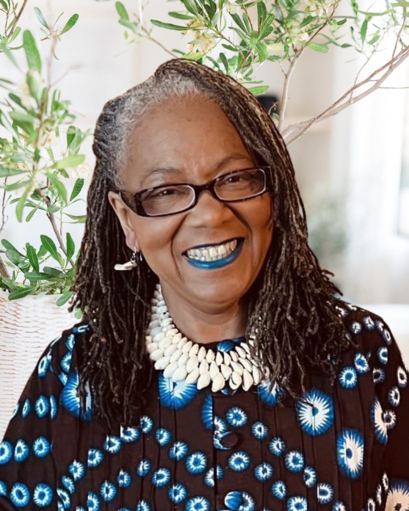 Paula Alphonse, smiling, wearing glasses and a white shell necklace